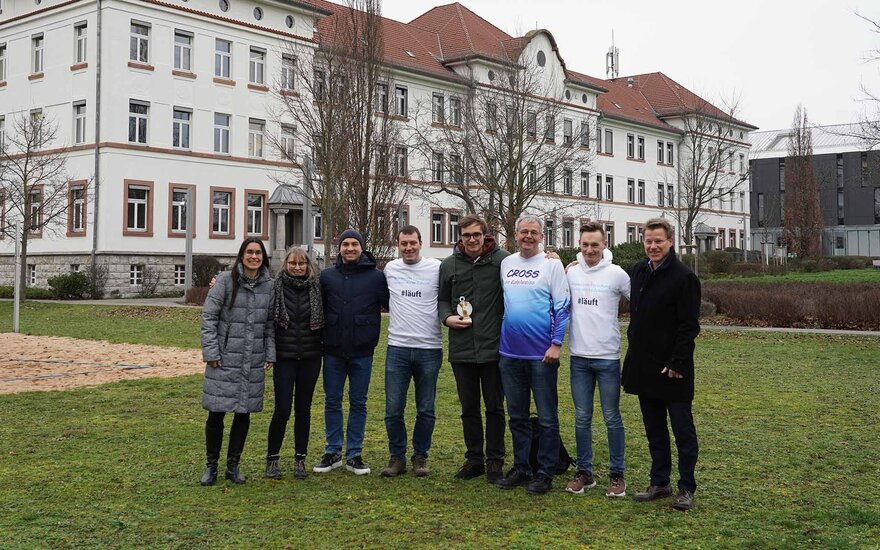 Acht Personen stehen Arm in Arm nebeneinander auf der Campuswiese vor einem weißen Hochschulgebäude