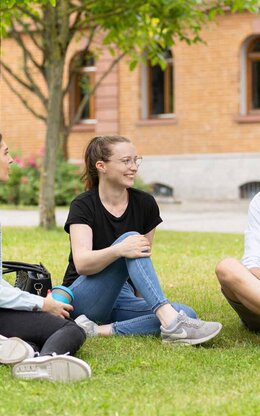 Drei Studierende sitzen auf der Campus Wiese und unterhalten sich