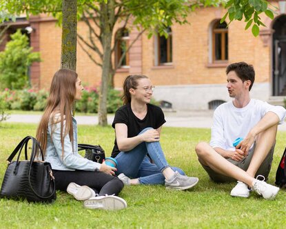 Drei Studierende sitzen auf der Campus Wiese und unterhalten sich