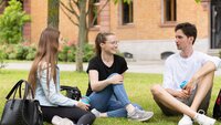 Drei Studierende sitzen auf der Campus Wiese und unterhalten sich