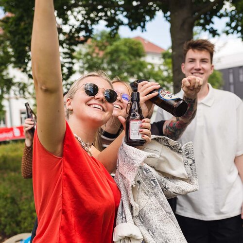 Drei Studierende feiern mit Bierflaschen auf dem Campus