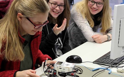 Schüler*innen sitzen an einem Schreibtisch und beschäftigen sich mit einem kleinen Roboter. 