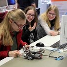 Schüler*innen sitzen an einem Schreibtisch und beschäftigen sich mit einem kleinen Roboter. 