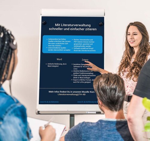 Drei Studierende hören einer Studierende, Referentin, Professorin, Dozentin zu, zeigt auf Flipchart mit Infos zum Zitieren