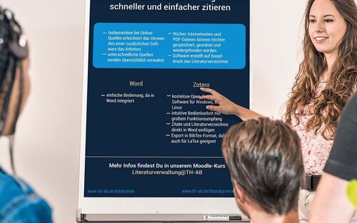 Drei Studierende hören einer Studierende, Referentin, Professorin, Dozentin zu, zeigt auf Flipchart mit Infos zum Zitieren