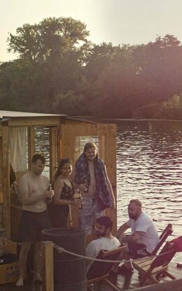 Sieben Studierende Gruppe auf hölzernem Hausboot am Steg auf dem Wasser bei Sonnenuntergang, gemütliches Beisammensein lachend und mit Flasche in der Hand