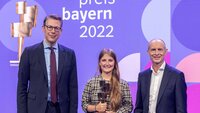 Nebeneinander vor einer violetten Wand mit der Aufschrift Kulturpreis Bayern 2022 steht ein Mann mit dunklem Anzug und Brille, eine junge Frau mit langen dunkelblonden Haaren und einer goldenen Statue in der Hand und ein Mann mit Glatze und ebenfalls dunklem Anzug