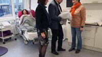 Ein Mann hält eine Babypuppe im Arm und steht zwischen zwei Frauen, die beide auf das Baby blicken. Im Hintergrund ist ein Bett mit der Simulationspuppe einer Frau zu sehen. Ringsherum sind weitere Einrichtungsgegenstände eines Kreißsaals zu sehen.