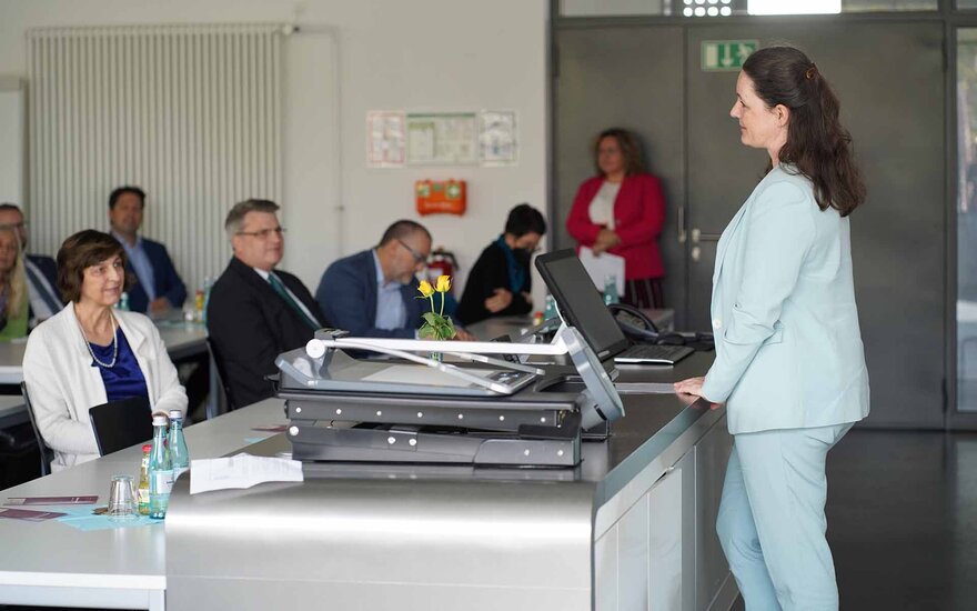 Eine Frau steht an einem breiten Tisch und spricht zum Publikum