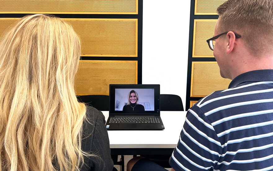 Eine Studentin und ein Student sitzen an einem Tisch. In der Mitte steht ein Laptop, auf dem gerade ein Meeting stattfindet.
