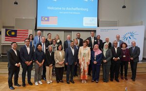 Mitglieder der Delegation aus Malaysia und Mitglieder der TH Aschaffenburg stehen als Gruppe in der Aula vor einer hellblauen Leinwand mit einem englischen Willkommensgruß, der malaysischen Flagge und dem TH-Logo darauf