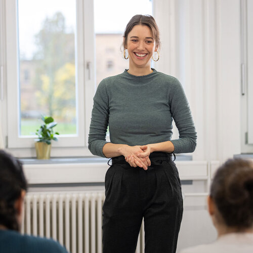 Eine junge Professorin steht zwei Personen bei einem Vortrag mit einem Lachen gegenüber. 