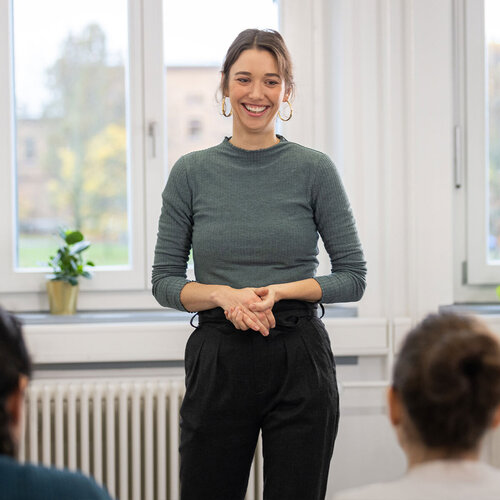 Eine junge Professorin steht zwei Personen bei einem Vortrag mit einem Lachen gegenüber. 