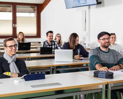 Studierende, die in mehreren Reihen hintereinander an Tischen in einem Hörsaal sitzen und lächelnd nach vorne blicken