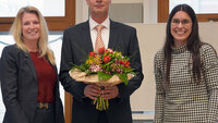 Der neue Vizepräsident steht in der Mitte auf der Bühne der Aula mit einem Blumenstrauß in der Hand, rechts neben ihm die Kanzlerin, links neben ihm die Hochschulratsvorsitzende