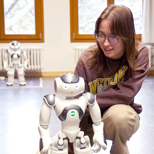 Mädchen schaut sich in der Hocke sitzend einen etwa 40 cm großen,  weißen Roboter an. Am anderen Ende des Raumes steht ein weiterer Roboter in der Unschärfe an einer Heizung.