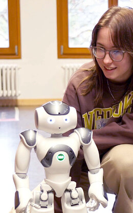 Mädchen schaut sich in der Hocke sitzend einen etwa 40 cm großen,  weißen Roboter an. Am anderen Ende des Raumes steht ein weiterer Roboter in der Unschärfe an einer Heizung.