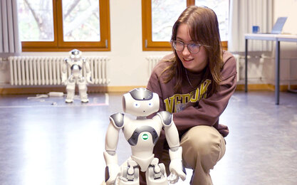 Mädchen schaut sich in der Hocke sitzend einen etwa 40 cm großen,  weißen Roboter an. Am anderen Ende des Raumes steht ein weiterer Roboter in der Unschärfe an einer Heizung.