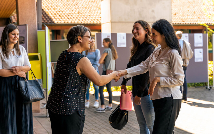 Zwei lächelnde Frauen geben sich zur Begrüßung die Hand, im Hintergrund weitere gut gelaunte Personen.