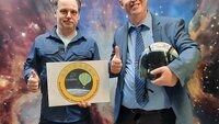 Professor Hildenbrand hält das Logo des Projekts in der Hand, Professor Döhring einen Astronautenhelm. Beide heben den Daumen der rechten Hand in die Höhe und stehen vor einer Wand, die einen Sternenhimmel zeigt