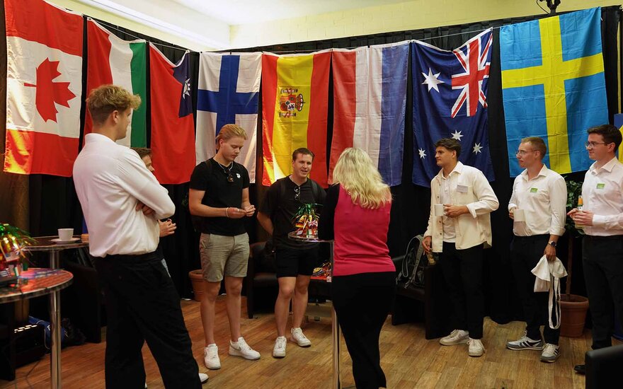 Junge Männer stehen in einem Raum, in dem viele unterschiedliche Länderflaggen hängen. In der Mitte steht eine Frau.