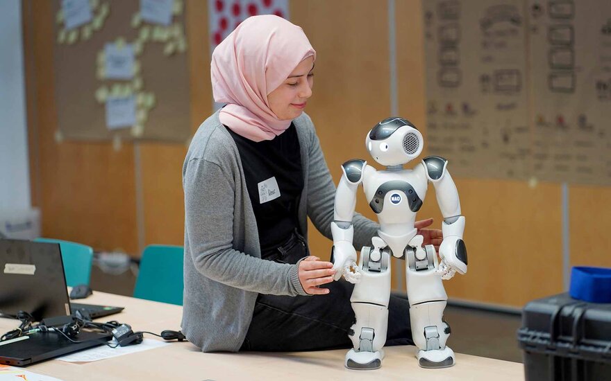 Ein Mädchen mit Kopftuch schaut den auf einem Tisch stehenden Roboter an