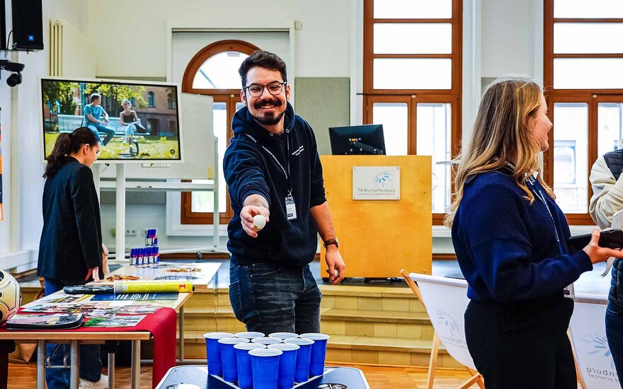 Ein Student zielt beim Bier-Pong in der Aula lachend mit einem Ball auf einen Becher.