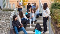 Ein Gruppe von internationalen Studierenden sitzt auf einer Treppe an der Hochschule vor einem gelbbraunen Backsteingebäude und ist im Austausch miteinander