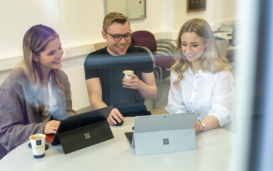 Luisa und zwei ihrer Kommilitonen sitzen vor zwei Laptops und unterhalten sich dabei lachend.