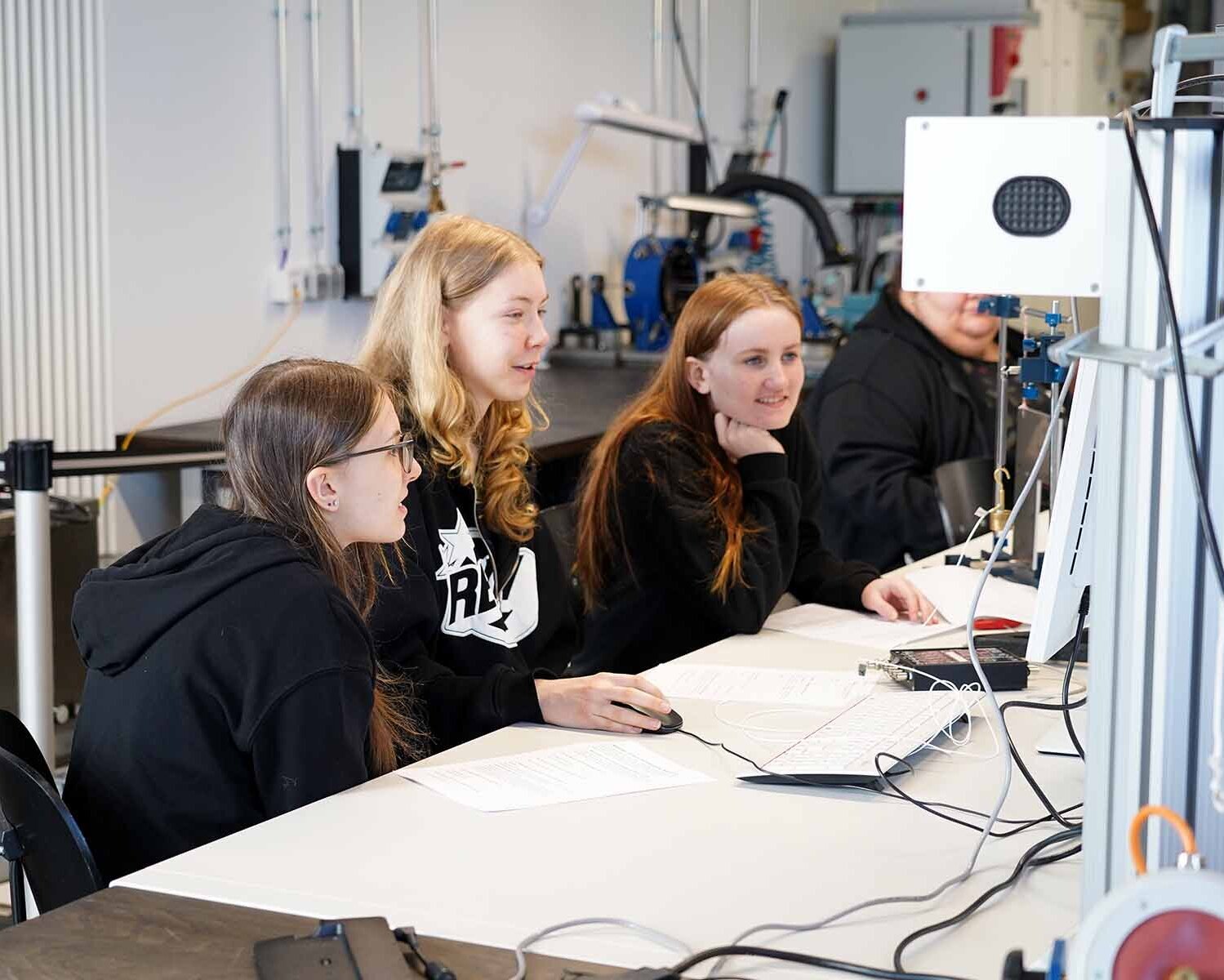 Drei Mädchen sitzen an einem Tisch vor einem Laboraufbau mit Computer