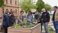 Eine Gruppe von Personen, in der Mitte die Präsidentin und die Kanzlerin stehen um ein Hochbeet mit Kräutern. Die Kanzlerin und die Präsidentin sowie die Nachhaltigkeitsbeauftragte pflanzen mit kleinen Schaufeln Kräuter ein.