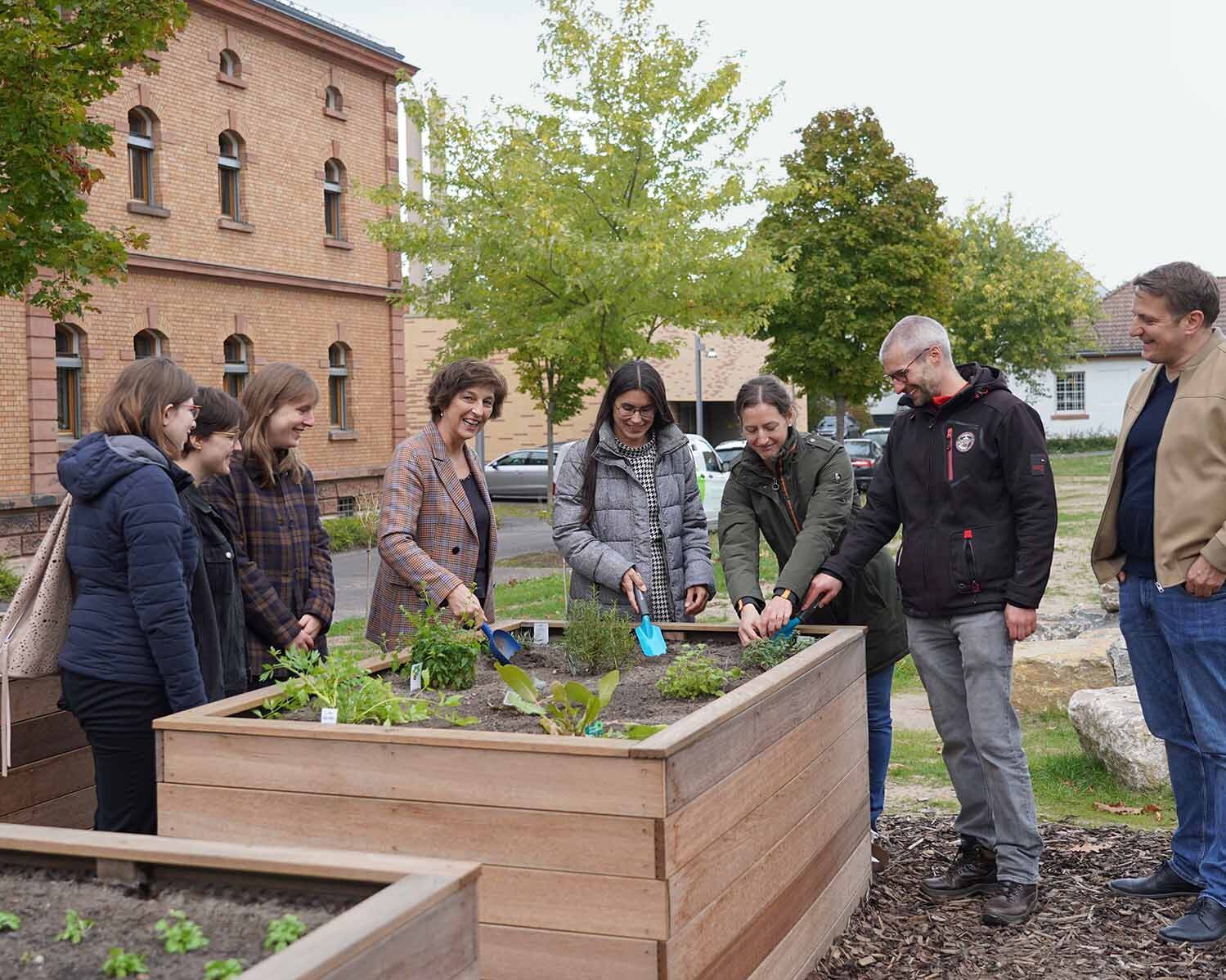 Eine Gruppe von Personen, in der Mitte die Präsidentin und die Kanzlerin stehen um ein Hochbeet mit Kräutern. Die Kanzlerin und die Präsidentin sowie die Nachhaltigkeitsbeauftragte pflanzen mit kleinen Schaufeln Kräuter ein.