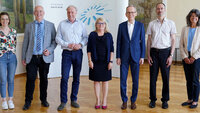 Eine Gruppe von sieben Personen stehen in einer Reihe lächelnd in einem Saal. Im Hintergrund das Logo der TH Aschaffenburg auf einer Messewand.