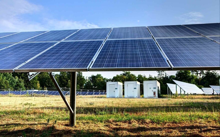 Solarpannels stehen auf einer grünen Wiese. Im Hintergund sieht man drei weiße Container.