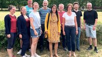 Gruppe von zehn Personen steht auf einer grünen Wiese, im Hintergrund grüne Bäume