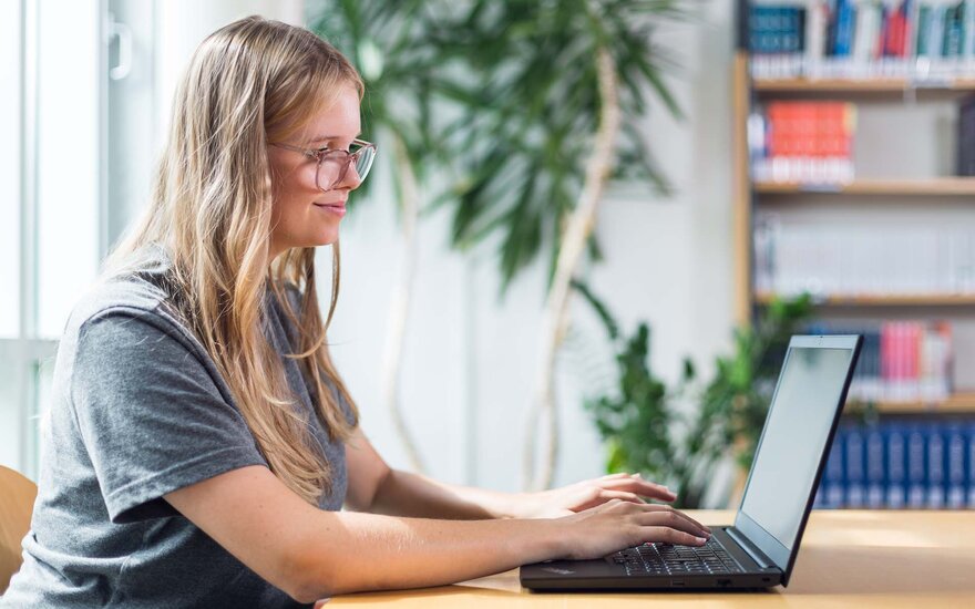 Blonde Frau sitzt an einem Tisch und arbeitet an einem schwarzen Laptop. Im Hintergund ist eine Pflanze und ein Bücherregal.