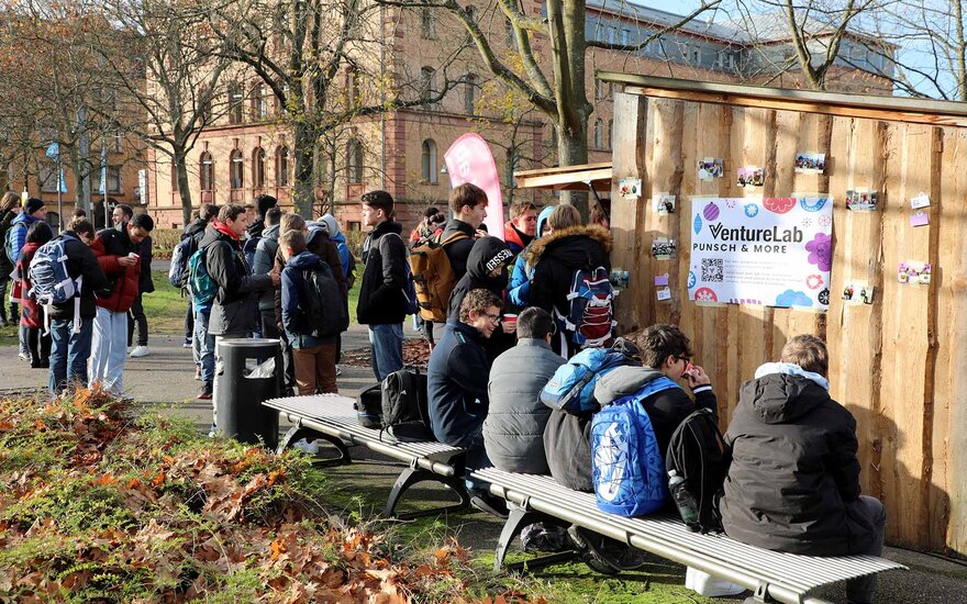 Eine Gruppe von Jungen und Mädchen steht vor einer Bude aus Holz auf dem Campus