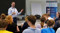 Projektleiter Professor Mewes spricht zum sitzenden Publikum im Hörsaal vor einer Tafel mit einem blau-weißen SANTO-Rollup rechts im Hintergrund