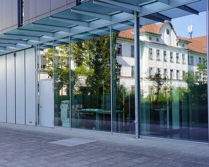 Weißes Gebäude der Hochschule mit rotem Ziegeldach spiegelt sich in der verglasten Fassade des modernen grauen Hochschulgebäudes