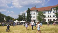 Campuswiese mit laufenden und spielenden Kindern und Erwachsenen und Ständen und Gebäude im Hintergrund