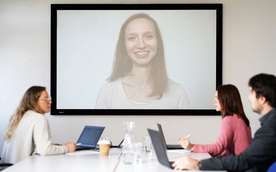 Mitarbeitende der Hochschule in Videokonferenz mit Übertragung von Frau, sitzend an Tisch mit Laptop und Unterlagen