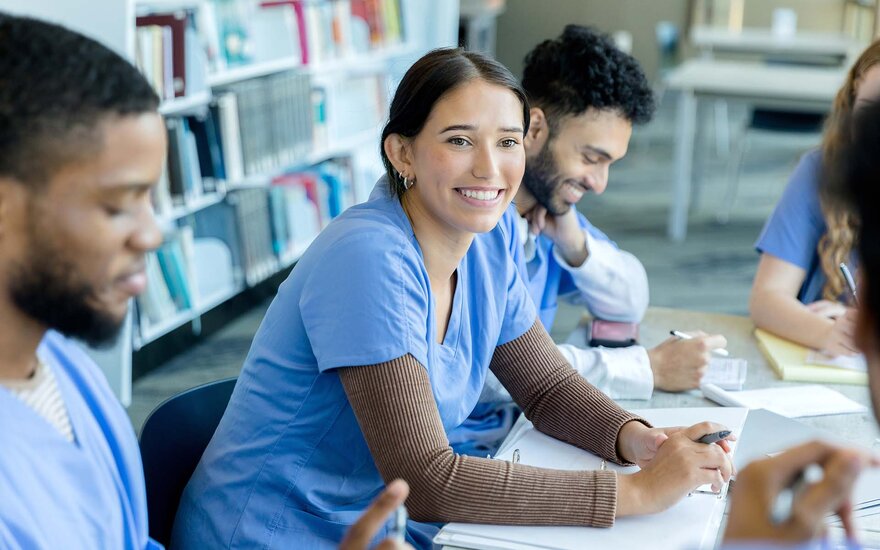 junge Frau sitzt mit Kommilitonen aus der Fakultät Gesundheitswissenschaften zusammen in der Bibliothek und lächelt.