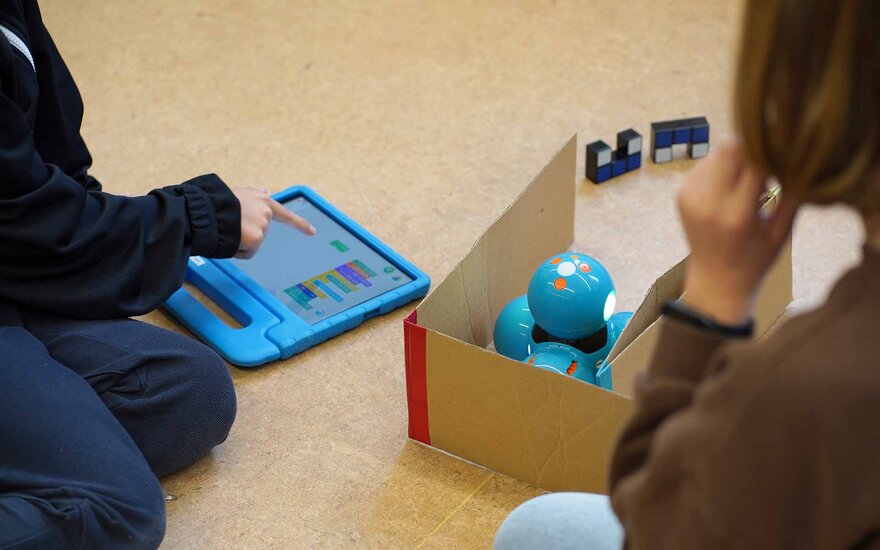 Kinder spilene mit einem Tablet und einem Mini Roboter auf dem Boden.