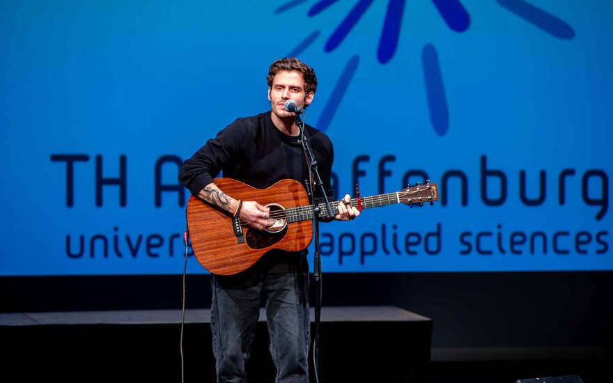 Musiker Chris Melon mit Gitarre auf der Bühne vor TH-Logo