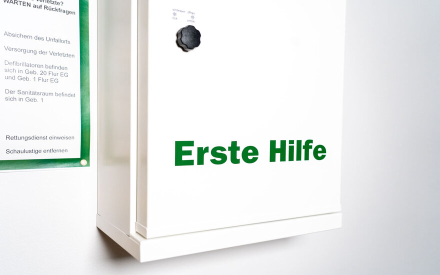 Ein weißer Kasten mit der grünen Aufschrift "Erste Hilfe" hängt an einer weißen Wand. Daneben hängt ein Dokument mit grüner Umrandung.