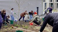 Mehrere Studierende lockern mit Rechen und Harken gemeinsam die Erde auf der Fläche vor dem Parkhaus auf, dessen graue Fassade im Hintergrund zu sehen ist.