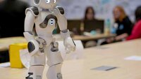 Menschenähnlicher Roboter steht auf einem Tisch im Hörsaal und im Hintergrund sind einige Schülerinnen bei der Gruppenarbeit zu sehen