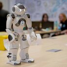 Menschenähnlicher Roboter steht auf einem Tisch im Hörsaal und im Hintergrund sind einige Schülerinnen bei der Gruppenarbeit zu sehen