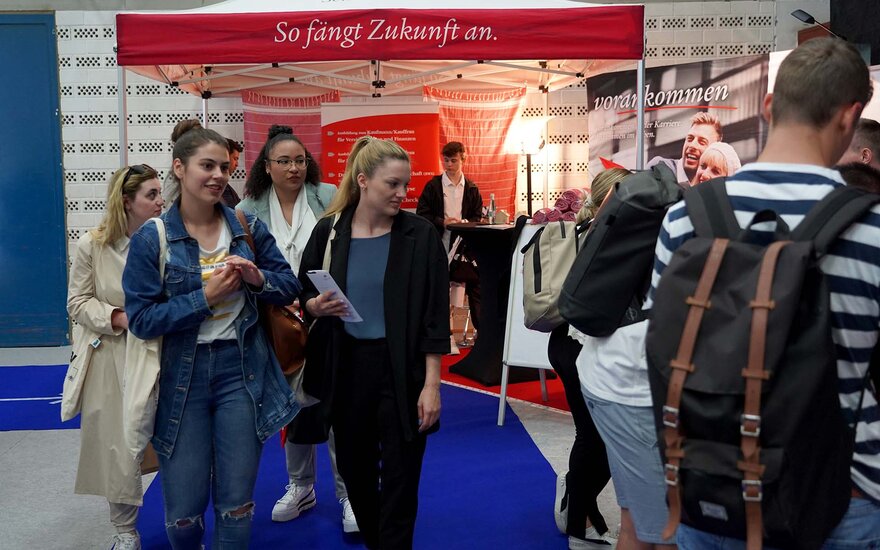 Eine Gruppe von Studierenden laufend vor einem Messestand und einige Studierende von hinten an einem Messestand