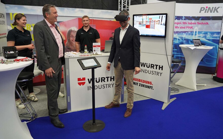 Zwei Männer stehen an einem Messestand, an dem weiterer Mann und eine Frau im Hintergrund stehen. Der rechte von ihnen hat eine schwarze VR-Brille auf.